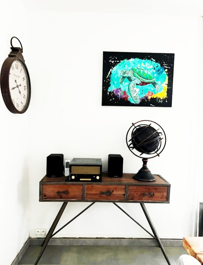 Photo d'une peinture tortue turquoise et colorée fond noir, mise en situation au mur au dessus d'un meuble bas avec des bibelots, mur blanc, horloge sur le mur.